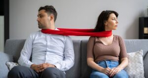 Couple showing codependent relationship signs, sitting apart on a sofa but tied together by a red scarf