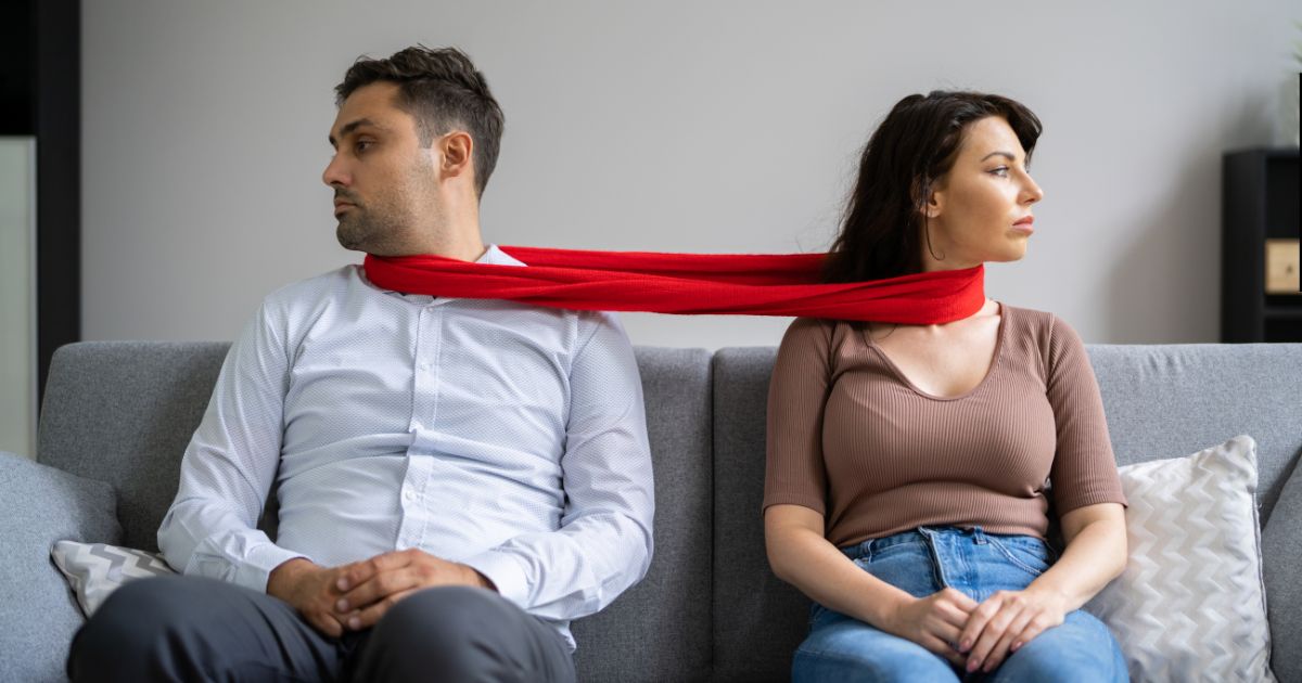 Couple showing codependent relationship signs, sitting apart on a sofa but tied together by a red scarf